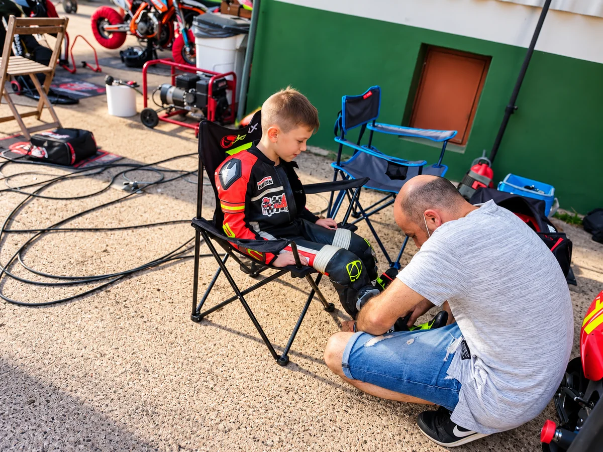 Un adulto ayuda a un joven piloto a prepararse en boxes junto a las motos