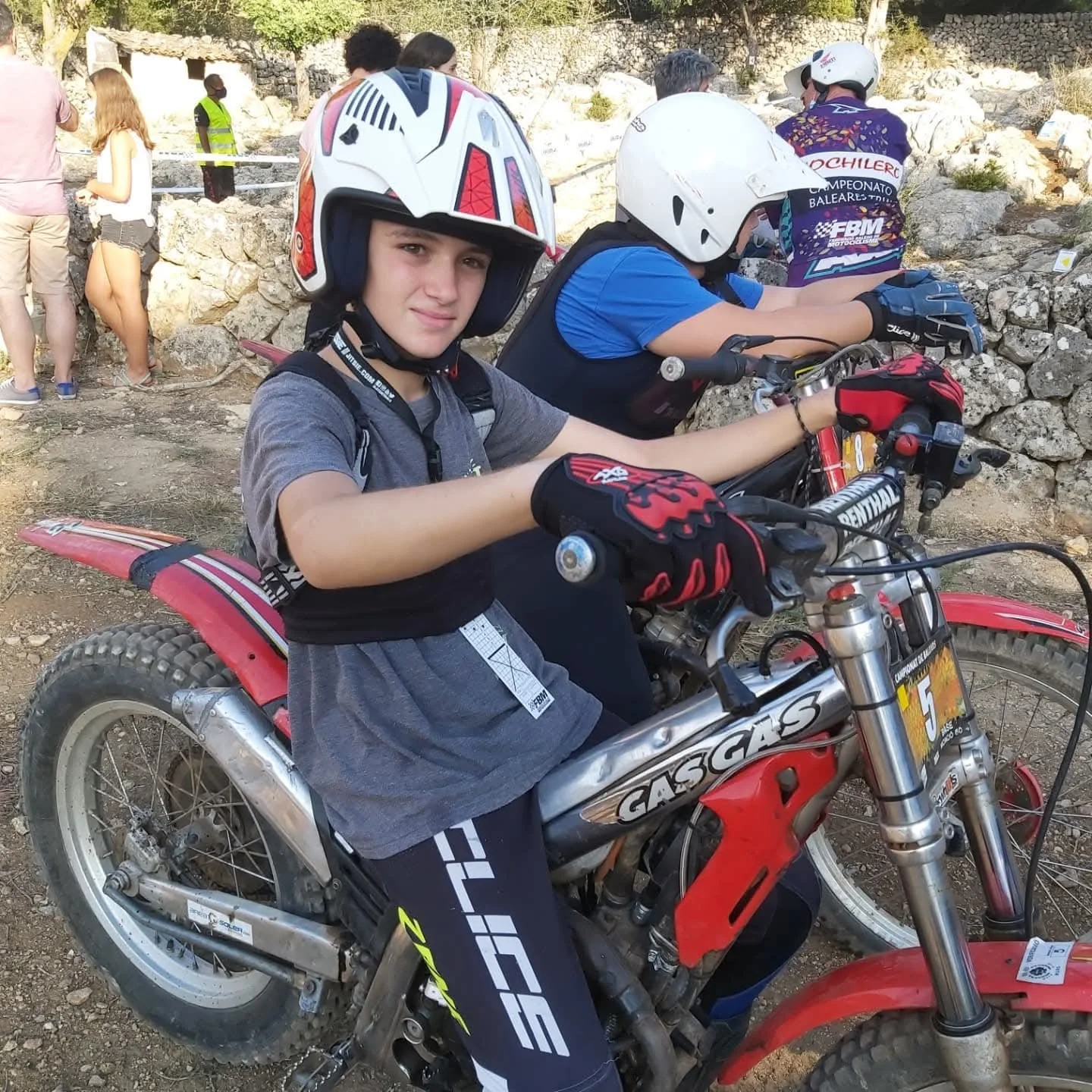 Dos pilotos con casco y protecciones junto a sus motos de trial, con una zona de obstáculos al fondo